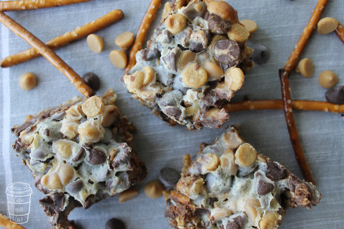 Three square Peanut Butter Pretzel Magic Bars over a grey linen background with pretzels and chocolate chips scattered about